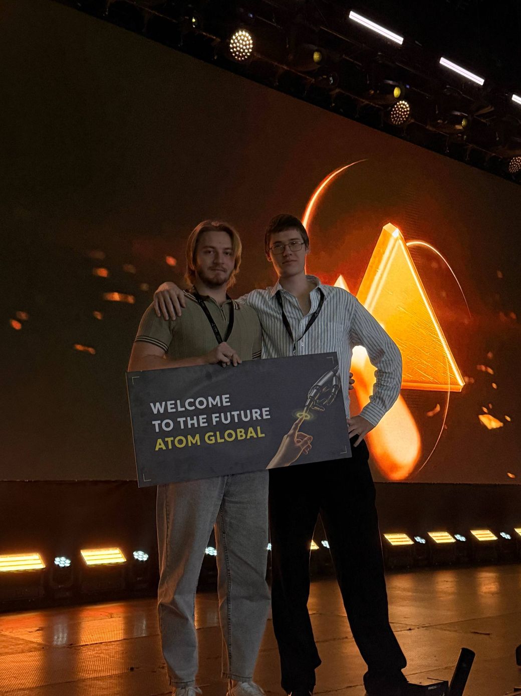 Two members of the MaxOut team on stage at Atom Global Expo, holding a sign that reads Welcome to the Future.